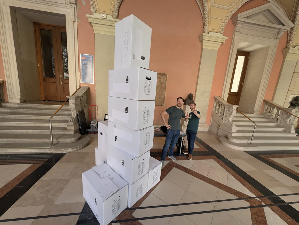 A tower of stacked white cardboard boxes, taller than the two people standing behind it, in a historic stone arcade with peach-colored walls.