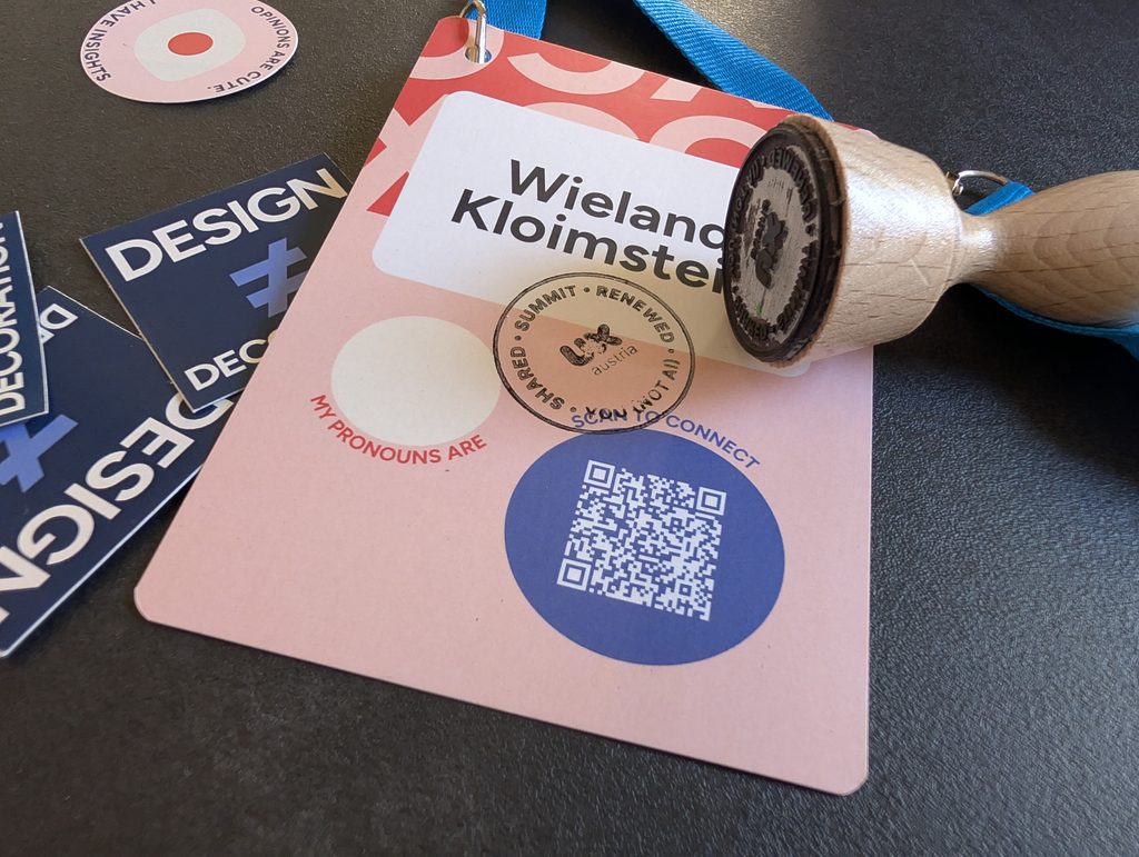 A pink conference badge on a table with a round stamp imprint on it, next to a wooden hand stamp and a blue lanyard.