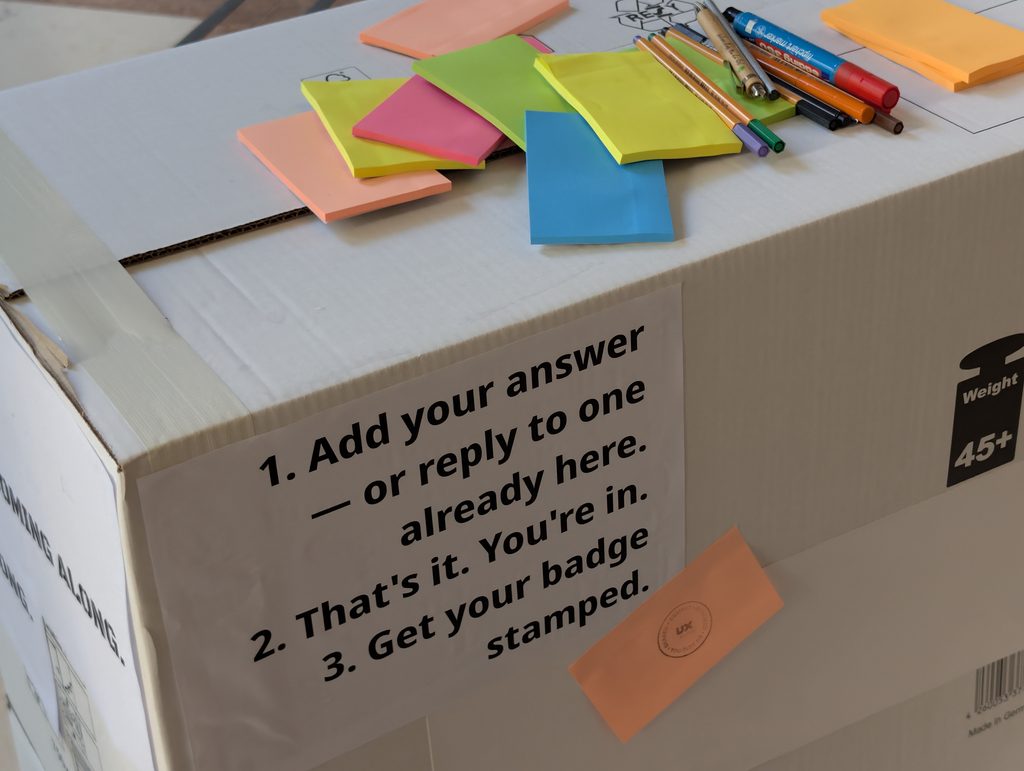 The top of a cardboard box with pens and colorful sticky notes; printed instructions on its side: add your answer or reply to one, then get your badge stamped.