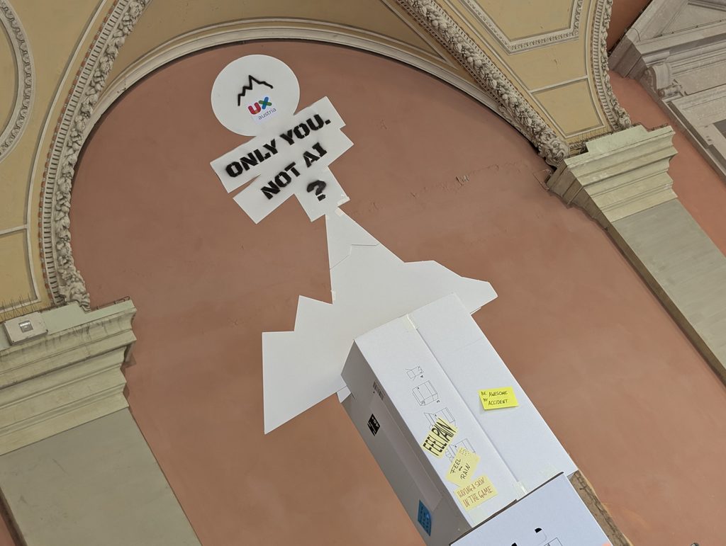 : Looking up at a white sign reading “ONLY YOU. NOT AI?” under a round UX Austria mountain logo, against an arched, painted ceiling.