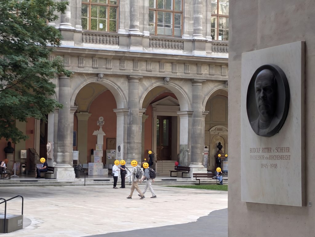 An arcaded university courtyard with a statue at its center and small groups of people walking, their faces covered by yellow smiley icons; a bronze portrait plaque at the right edge. Fare away: the cardboard tower