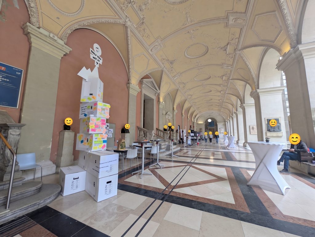 A long stone arcade with the sticky-note-covered tower in the middle distance, standing tables around it and a few people, their faces covered by smiley icons.