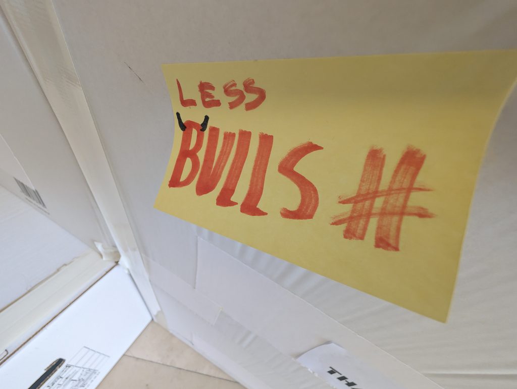 Close-up from below of a yellow sticky note with “LESS BULLSH#T” handwritten in red marker, against a white ceiling.
