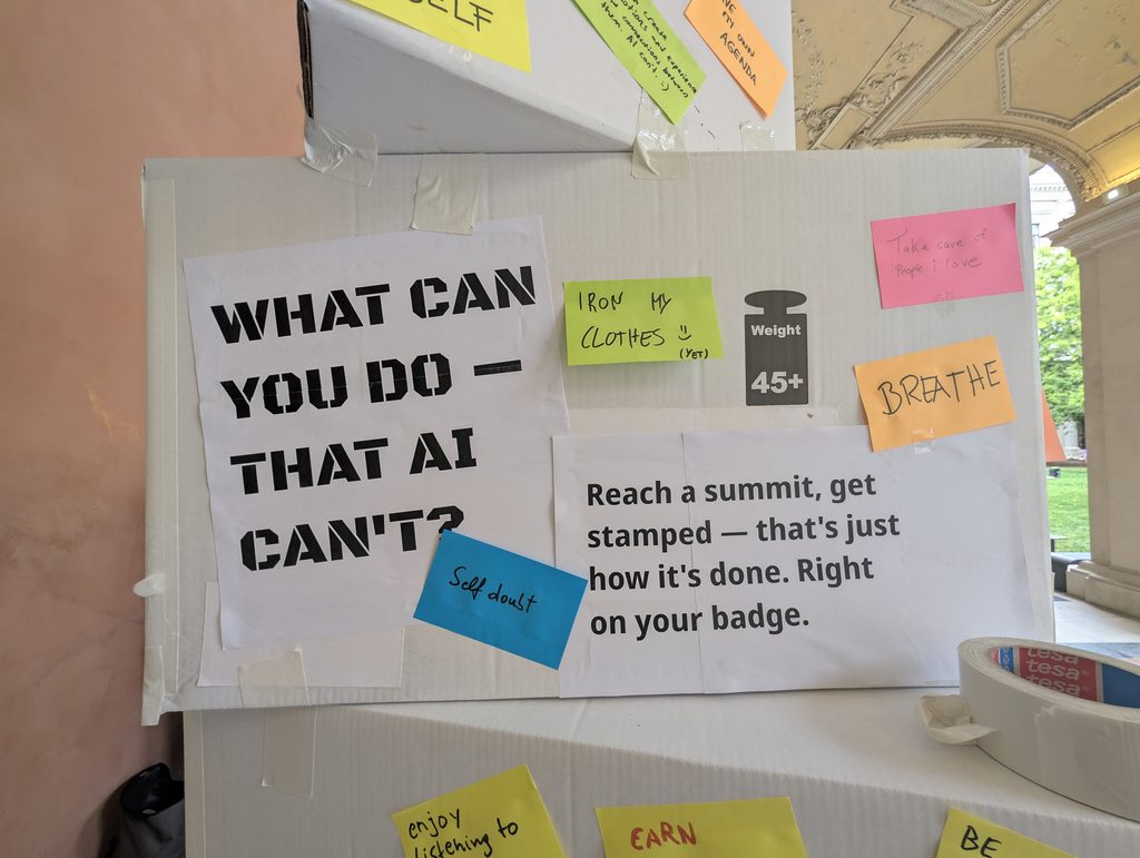 The tower's main sign “WHAT CAN YOU DO — THAT AI CAN'T?” beside a printed note “Reach a summit, get stamped”, with notes “IRON MY CLOTHES (YET)”, “BREATHE” and “Self doubt”.