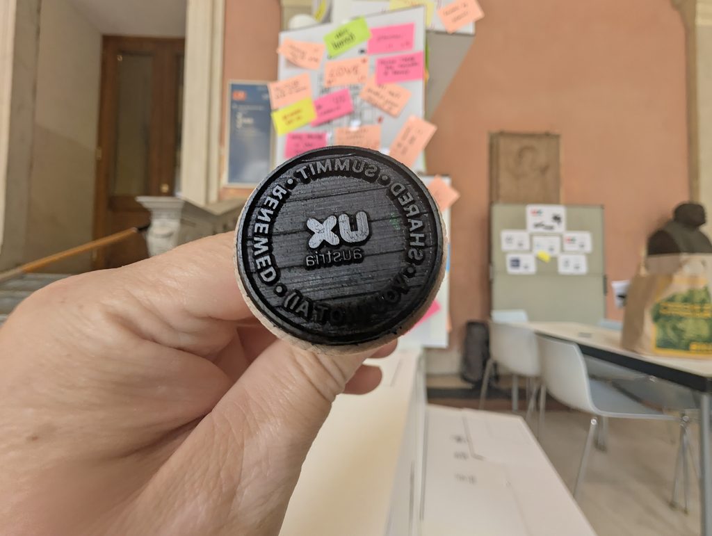 A hand holds up a round black stamp; its face shows mirrored lettering (“Summit”, “Renewed”, “Shared”) around a UX Austria logo, with the tower blurred behind.