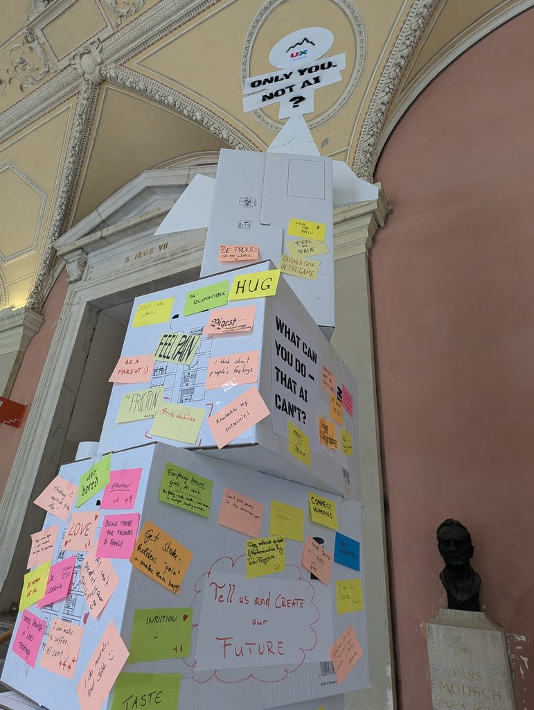 Looking up at the top of the tower: the sign “ONLY YOU. NOT AI?” above stacked boxes and a board covered with sticky notes, including “HUG”.