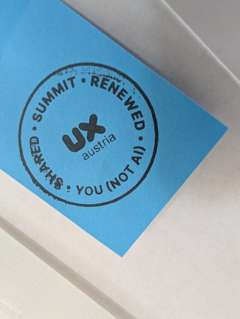 A blue sticky note with a round black stamp imprint reading “Shared · Summit · Renewed” and “You (not AI)” around a UX Austria logo.