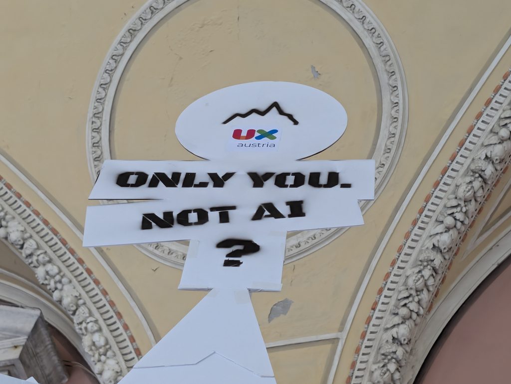 Close-up of the “ONLY YOU. NOT AI?” sign with the round UX Austria mountain logo above it, against an ornate ceiling.