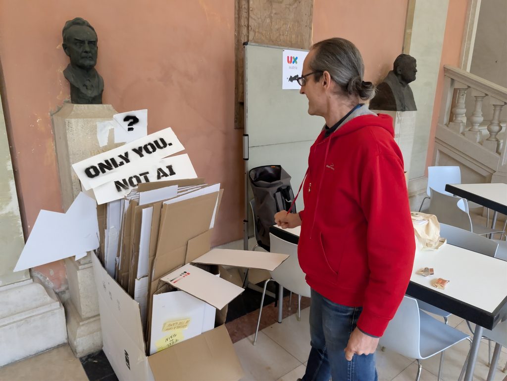 Taking down the tower: a man in a red hoodie looks toward the table, next to flattened cardboard and stencilled cardboard signs reading “ONLY YOU. NOT AI?”, between two bronze busts in the arcade.